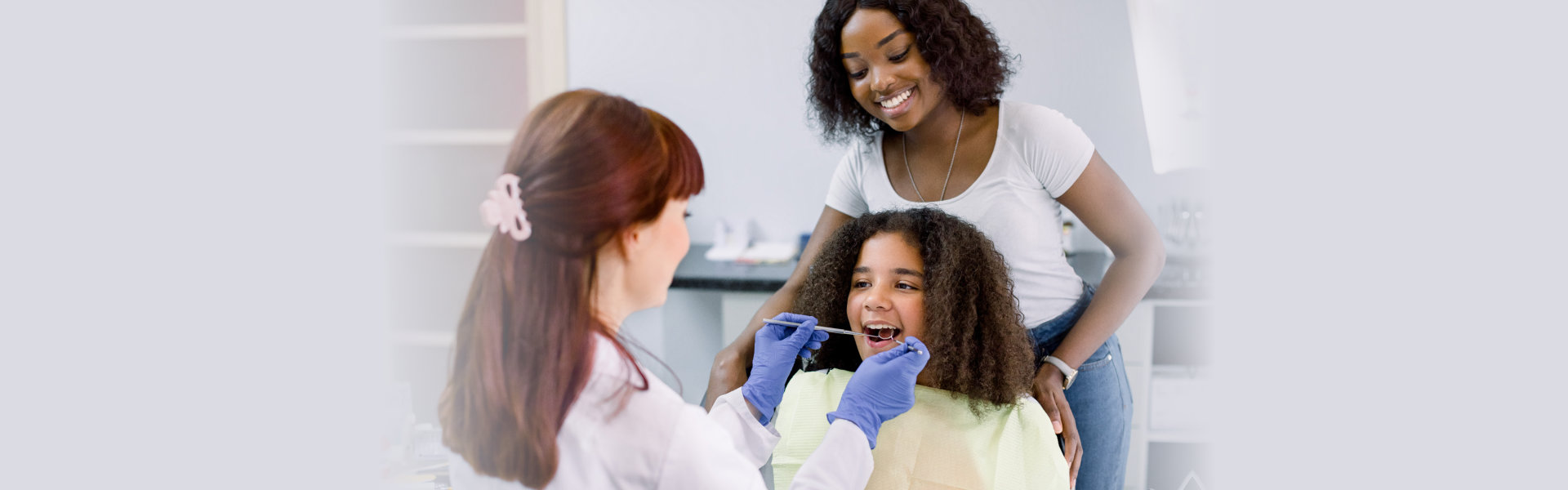 Dental treatment without fear. Female dentist in white uniform and gloves, examining teeth of little African American girl, while her mother supporting her behind, at modern pediatric dental clinic